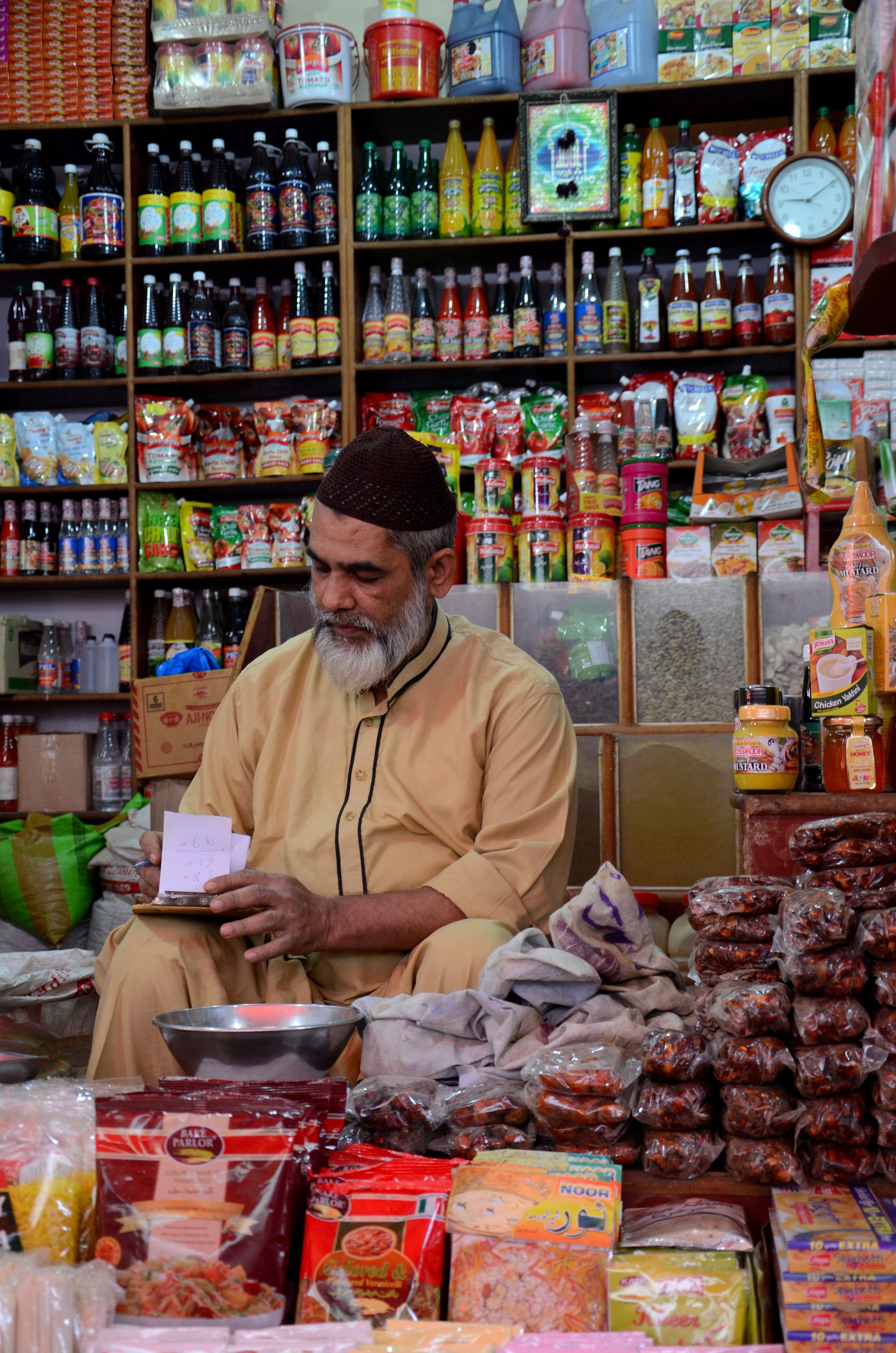 A shop owner tends to paperwork while sitting inside his general provision store at the historical Empress Market in Karachi, Pakistan; with a colourful display